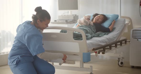 Smiling Afroamerican Nurse Adjusting Hospital Bed for Preteen Asian Girl alt