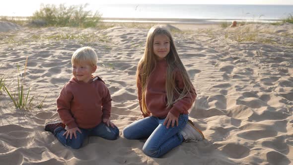 Brother and Sister in a Hoodie Sit on the Sand alt