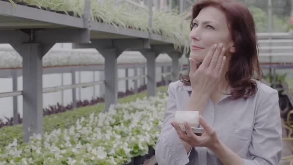 Beautiful Mid-adult Caucasian Woman Applying Organic Moisturizer in Greenhouse. Portrait of Positive alt