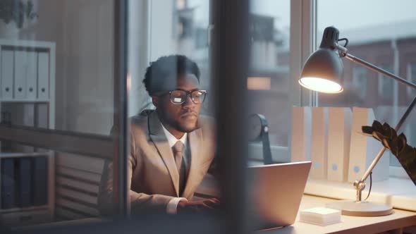 Afro-American Male Clerk Working on Laptop at Office Desk in the Evening alt