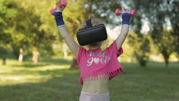 Athletic Child Girl in VR Headset Helmet Making Fitness Workout Exercises with Dumbbells in Park alt