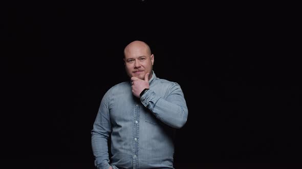 A bald man strokes his beard and poses for the camera on a black background alt