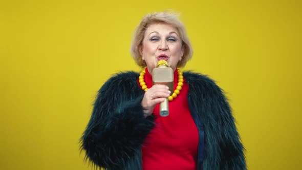 Portrait of Elegant Senior Woman Singing on Microphone on Yellow Background alt