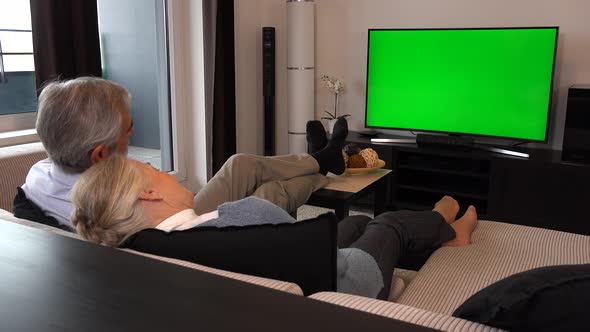 An Elderly Couple Sits in A Living Room in An Apartment and Watches TV with A Green Screen alt