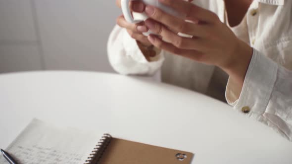 Young Pensive Girl In A White Shirt Drinking Morning Coffee While Sitting At A White Table, Planning alt