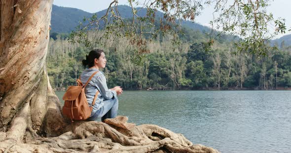 Woman sit on the tree root and enjoy the view of the lake alt