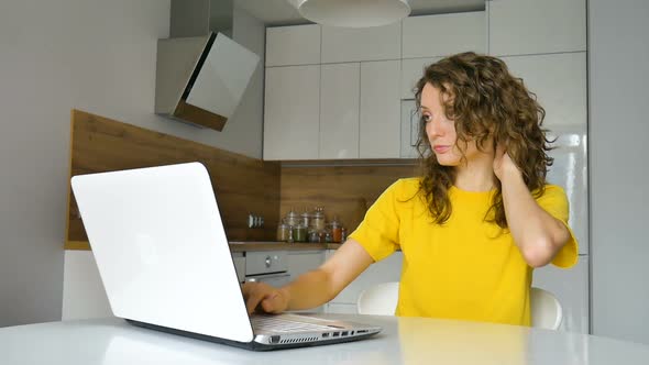 Young Woman with Curly Hair and Yellow Shirt is Working From Home Using Her Laptop at the Kitchen alt