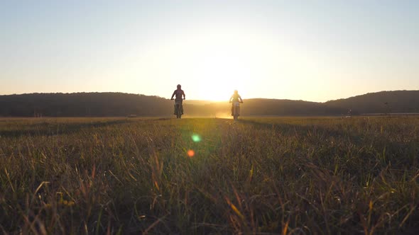 Two Motorcyclists Passing Through Large Field with Beautiful Sunset at Background alt