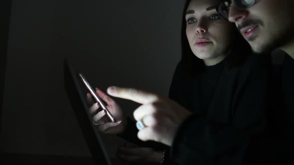Young woman and young woman indoor in apartment at darkness using computer and smartphone alt