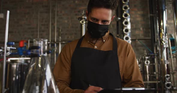 Portrait of caucasian man working at gin distillery wearing face mask and apron looking to camera alt