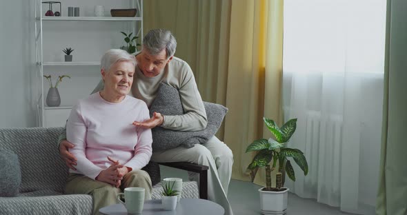 Elderly Caucasian Couple Sits on Gray Sofa Old Man Loving Husband Hugs His Wife Comforts Beloved Sad alt