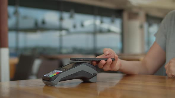 Female hand paying bill with mobile phone and contactless terminal staying on the cafe table alt