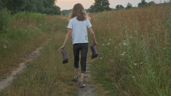 Girl Walking on Rural Road with Boots in Her Hands alt