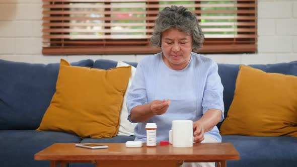 Asian senior elderly woman taking pills, drinking water in the living room at home.