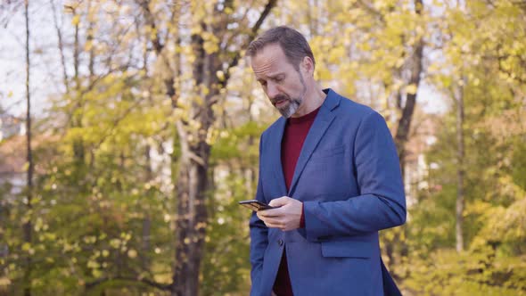 A Middleaged Handsome Caucasian Man Works on a Smartphone and Walks in a Park in Fall