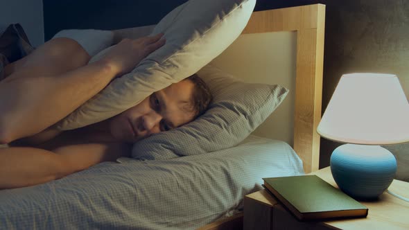 Man Cannot Fall Asleep Lying in Bed at Night Due to Noise Covers His Head with a Pillow alt