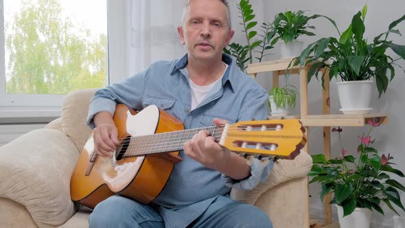 Senior Man Plays Acoustic Guitar and Sings in Apartment Living Room alt