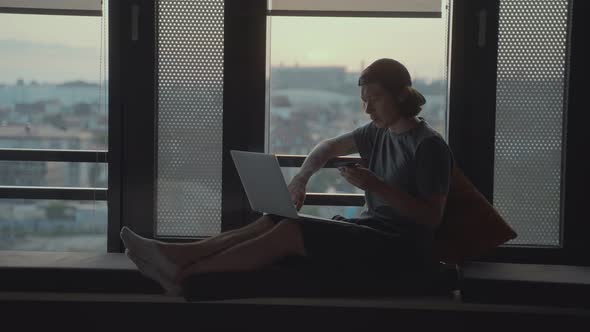 Handsome Hipster Man Sits Alone in Front of a Panoramic Window with a Laptop