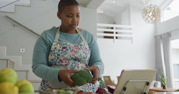 Happy african american plus size woman wearing apron, cooking, using tablet in kitchen alt