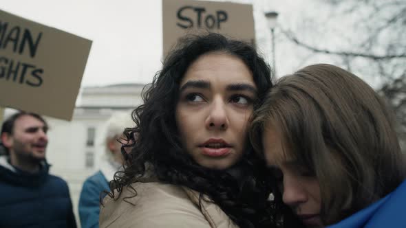Two embracing woman covered with flags and group of young caucasian people manifesting against war i alt
