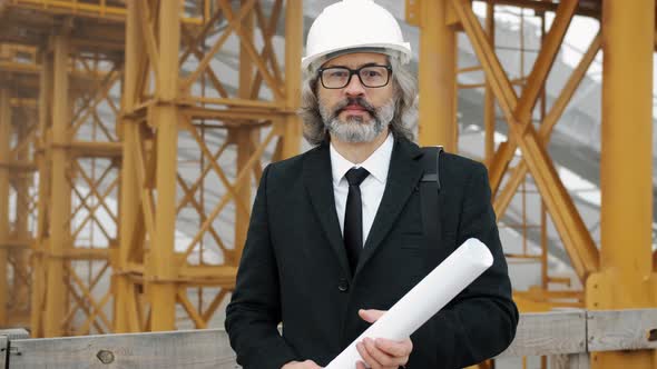 Slow Motion Portrait of Senior Engineer Standing in Building Area Holding Blueprint Touching Glasses alt