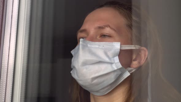 A young woman in a medical protective mask looks out the window alt