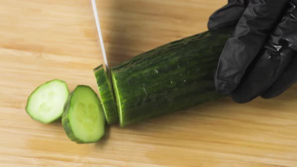 Chef's Hands in Black Gloves Cutting Fresh Cucumber Into Slices on Wooden Board