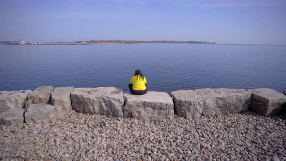 Person in Toronto Trillium Park with headphones sitting near lake in bright yellow jacket during day alt
