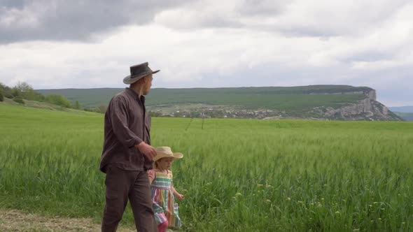 A Farmer Father with Happy Small Daughter Inspects a Green Wheat Field in Spring alt