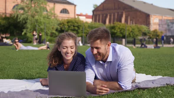 Young Couple Lying on Lawn and Watching Laptop Content on Beautiful Sunny Day alt
