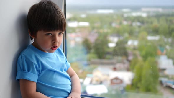 Child Boy in a Blue T-shirt Sits on the Window of a High Floor alt
