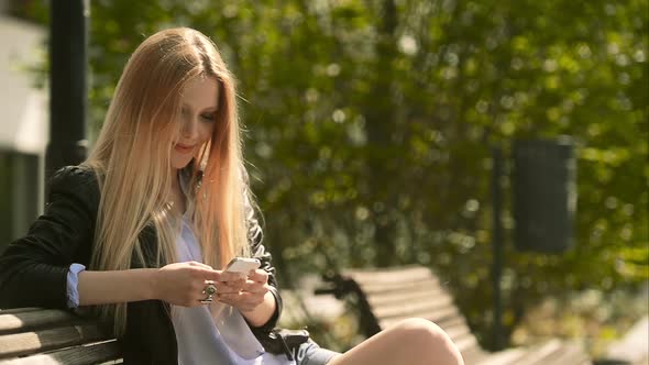 Young Beautiful Blonde Girl in Summer Day is Sending Messages with Smartphone Sit on a Bech at Urban