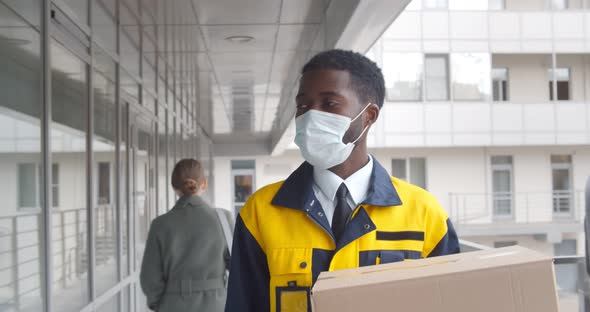 Afroamerican Courier in Safety Mask Delivery Package in Modern Office Building alt