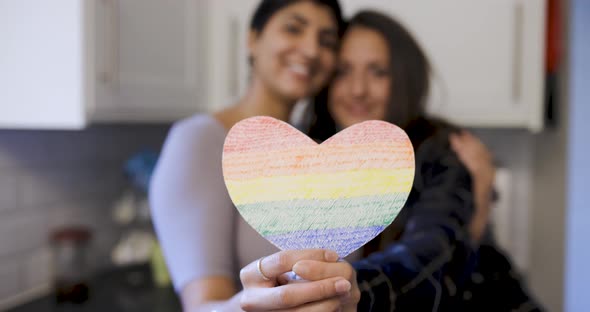 Happy girl friends lesbian couple embracing and holding a heart shaped rainbow flag alt