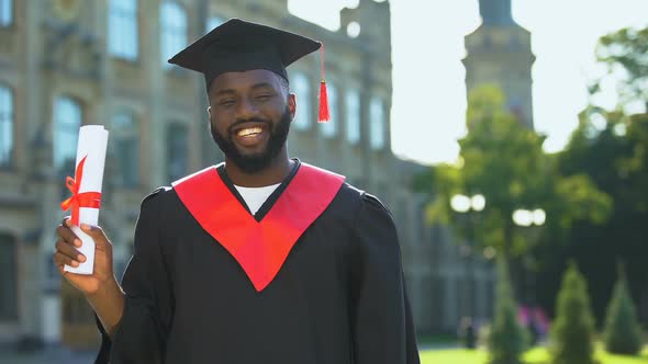 Excited Afro-American Student in Graduation Gown Holding Diploma Looking Camera alt