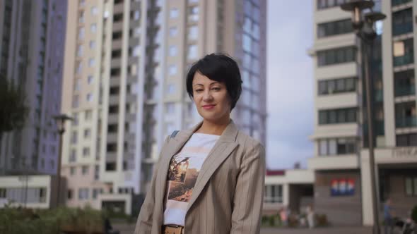 Brunette Stylish Woman Stands in the City Near Houses and Smiles alt