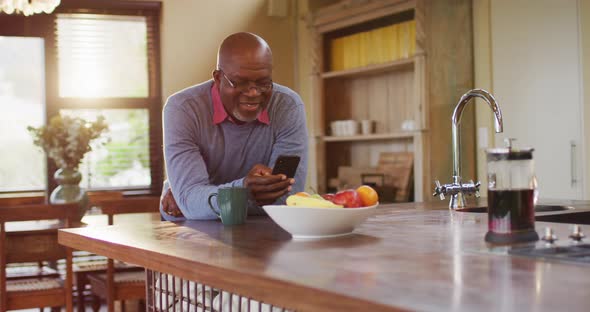 African american senior man leaning on kitchen counter using smartphone, taking off glasses, smiling alt