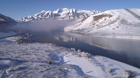 Flying towards snow covered mountain over lake with low foggy clouds alt