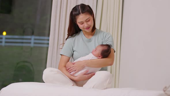 Beautiful Asian mom woman holding newborn baby in her arms sit on bed at white cozy home alt