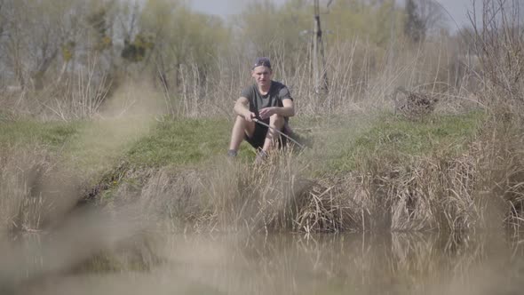 Wide Shot Portrait of Focused Young Caucasian Fisherman Sitting on Bushy Lake Shore and Looking at alt