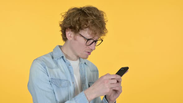 Redhead Young Man Loss on Smartphone, Yellow Background  alt