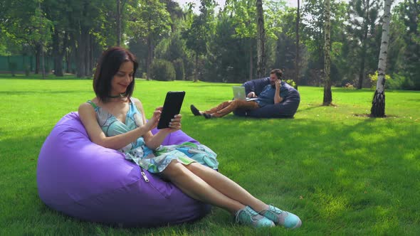 Young Brunette Girl Is Sitting in a Chair in the Park and Swipe Pages on Your Tablet alt