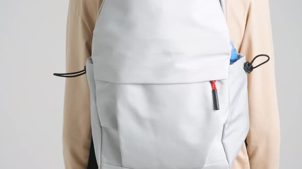 Man in light clothes with gray backpack on his back walks away from camera on white background