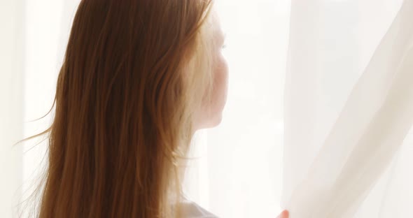 Young woman opening curtain looking through window in the morning at home her. alt