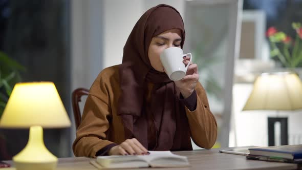 Serious Concentrated Young Muslim Woman in Hijab Reading Book in the Evening at Home alt