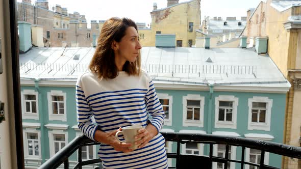 Young Woman Drinking Hot Coffee on Balcony and Observing Cityscape in Downtown.