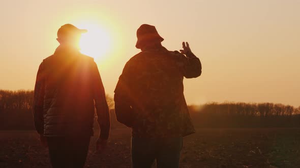 An Elderly and Young Farmer Go Together Over a Plowed Field at Sunset alt