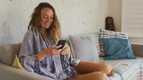 Smiling caucasian woman wearing blanket, sitting on sofa using smartphone in cottage living room alt