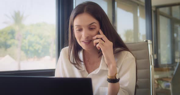 A businesswoman talking on the phone, smiling, while working on a laptop at the office alt
