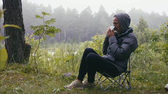 Woman hiker in jacket drinking tea in forest sitting on chair in nature camping.
